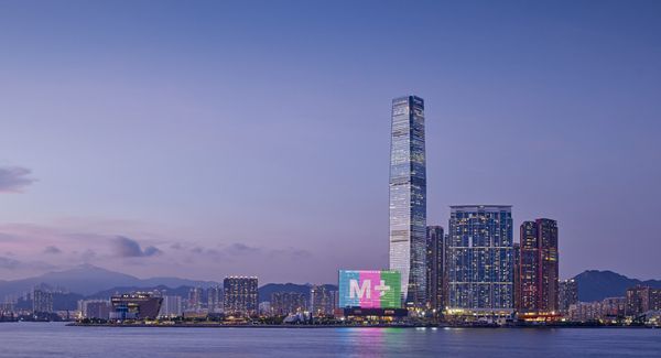 照片展示暮色下的香港维多利亚港，对岸是高楼大厦林立，遍布镜头下方。位于观者右方的楼群巍然屹立，当中包括一座长方形的建筑，外墙亮着“M+”的字样。