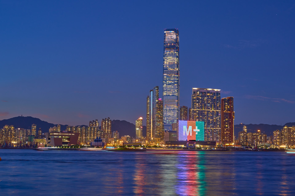 View of buildings across Hong Kong's Victoria Harbour at dusk, which stretches across the frame at the bottom. A campus of buildings stands on the viewer's right, including a rectangular block illuminated with the words 'M+'.